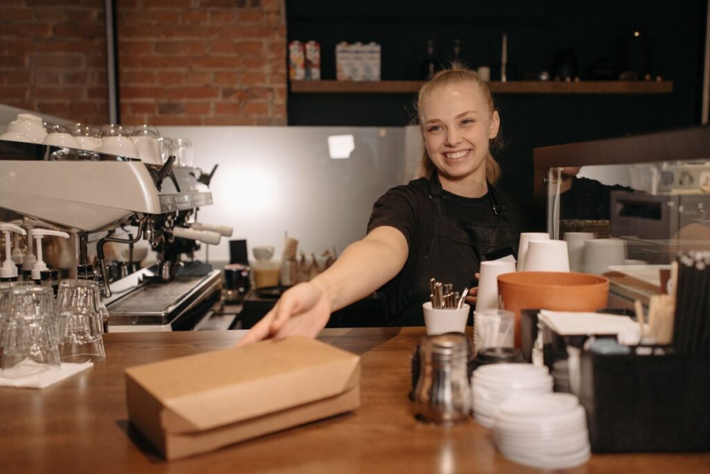 Smiling female barista handing a takeout box at a modern coffee shop counter.